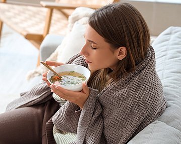 Suppe ist ein wohltuendes Hausrezept gegen Erkältung mit Schnupfen. Frau mit Decke um den Schultern, sitzt auf der Couch und isst Suppe.