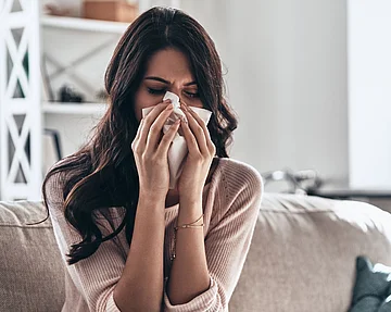 Tipps gegen Schnupfen können helfen, einer Nasennebenhöhlenentzündung vorzubeugen. Frau mit Schnupfen schnäuzt in ein Taschentuch.
